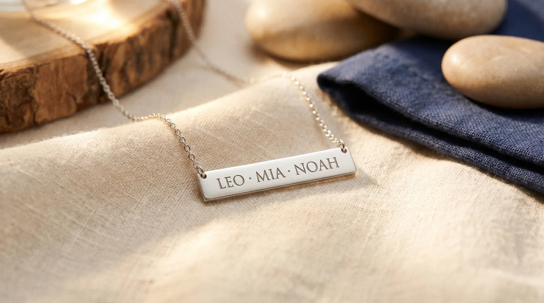A horizontal bar pendant engraved with three children's names resting on warm cream linen