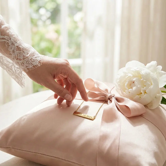 Bride's hand reaching toward a personalized engraved date necklace on a satin pillow with a white peony